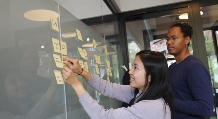 Two individuals writing on sticky notes on a glass wall, collaborating on knowledge management strategies.