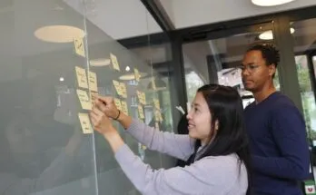 Two individuals writing on sticky notes on a glass wall, collaborating on knowledge management strategies.