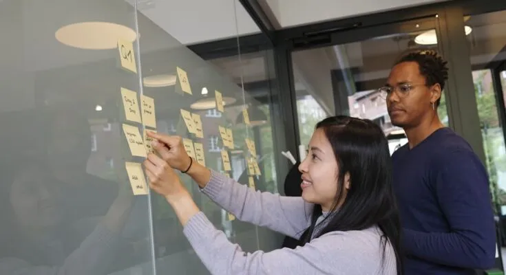 Two individuals writing on sticky notes on a glass wall, collaborating on knowledge management strategies.